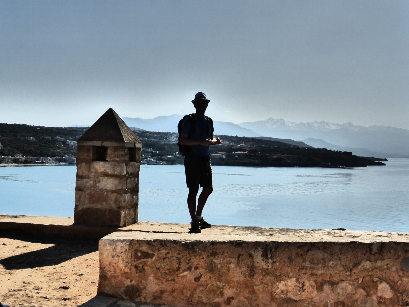 Silhouette of a hiker on a stone rampart, facing a blue bay and snow-capped mountains