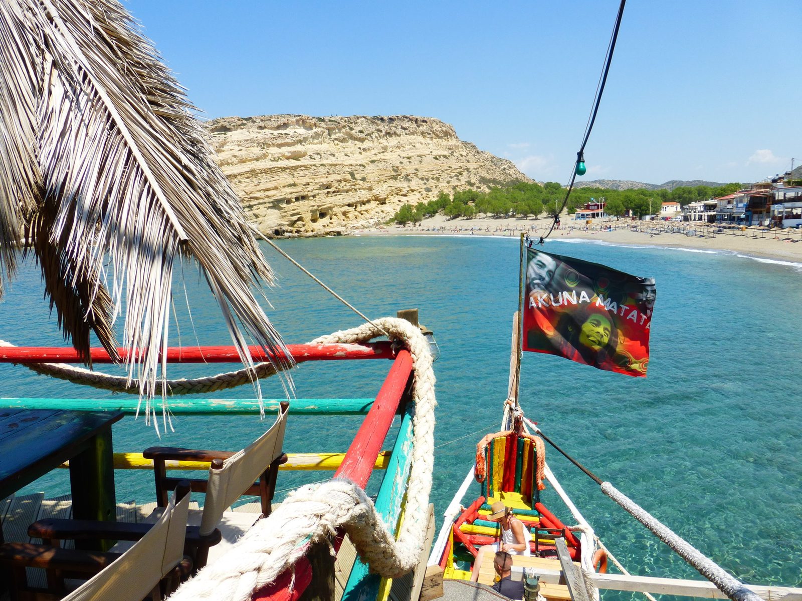 Matala beach seen from a brightly colored beach bar: turquoise sea, sandstone cliff and a “Hakuna Matata” flag