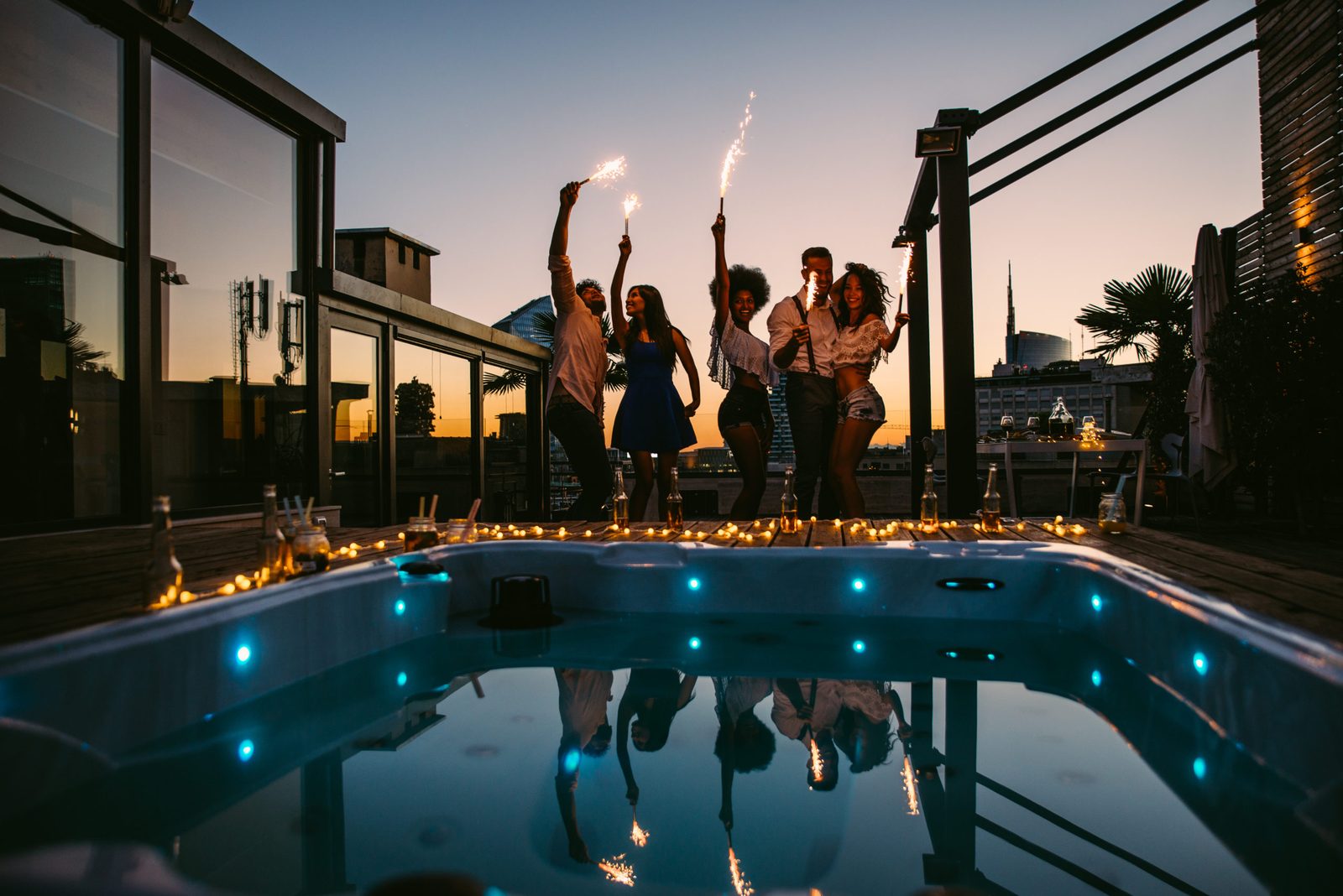 Group of friends waving sparklers on a rooftop terrace at dusk, next to a lit-up hot tub