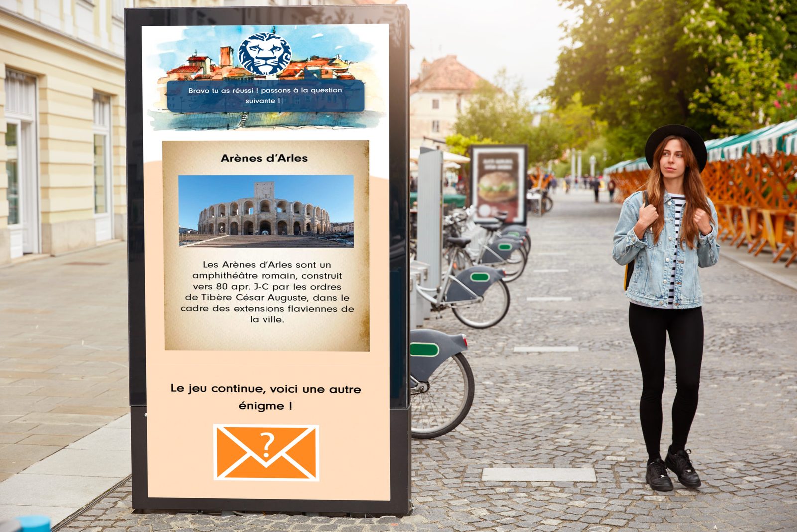 Display kiosk in a pedestrian street showing the game: an “Arles Amphitheatre” card and “The game goes on, here’s another riddle!”, next to a tourist in a black hat