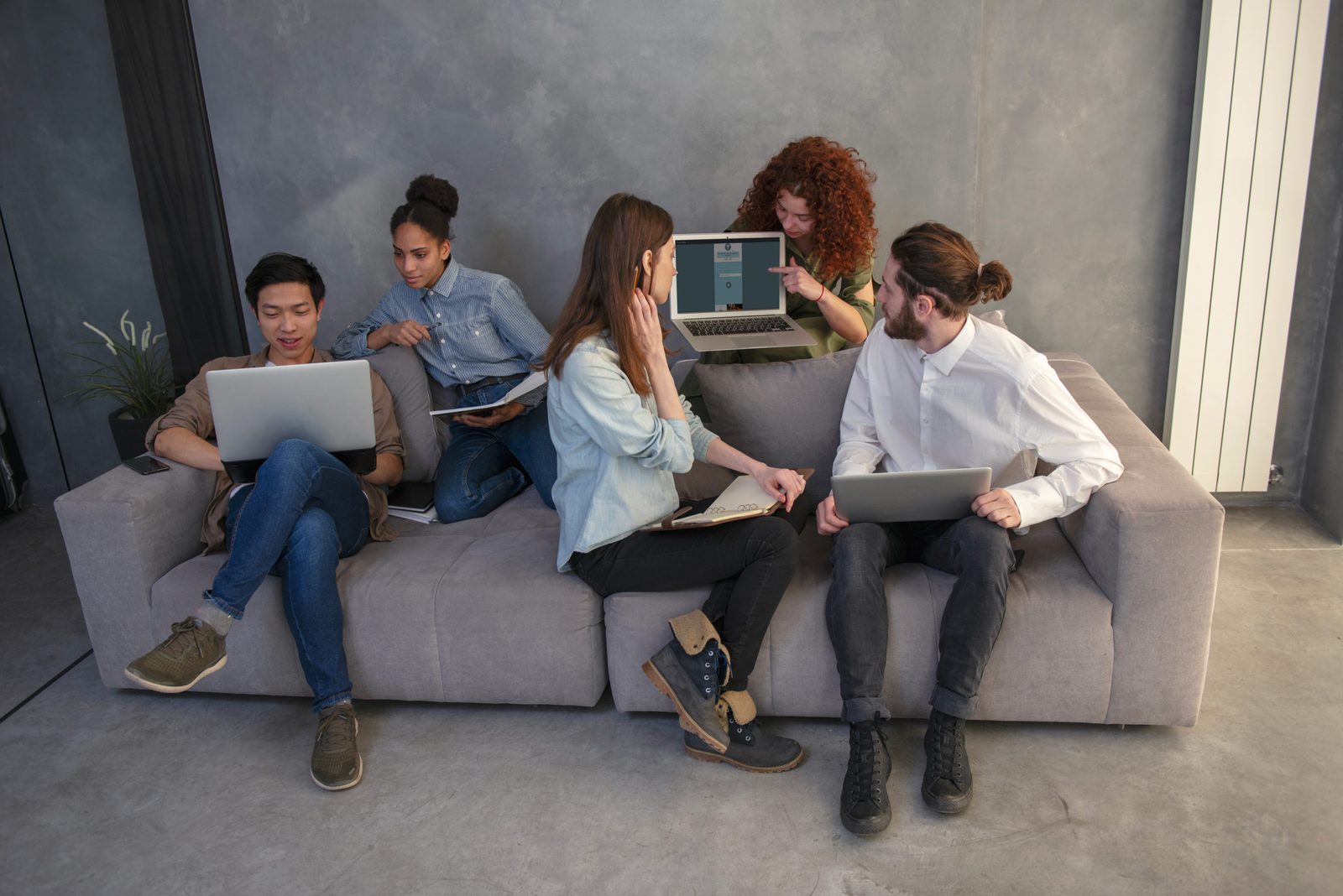 Five young colleagues sitting on a sofa with their laptops, one of them showing her screen to the others